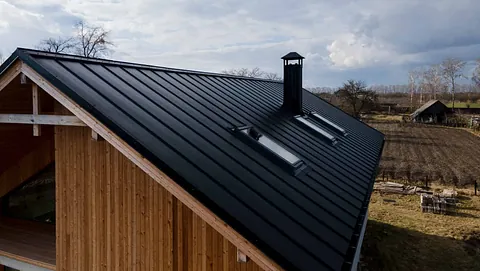 Moderne hus med svart ståltak, to takvinduer og pipe, omgitt av jorder og trær under overskyet himmel.